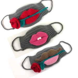 May include: Three gray crocheted face masks with different designs. The first mask has a red rose and red lips with a green stem. The second mask has pink lips with white teeth and a green stem. The third mask has brown lips with a pink rose and a green stem.