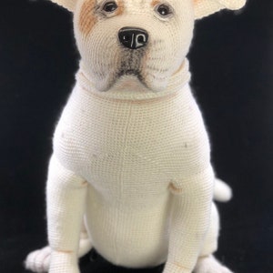 May include: A white crocheted stuffed dog with brown markings around the eyes. The dog is sitting with its paws tucked underneath it.