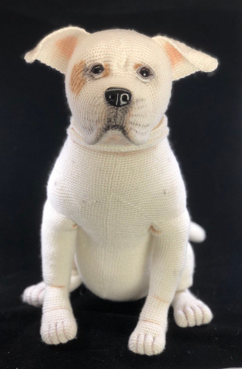 May include: A white crocheted stuffed dog with brown markings around the eyes. The dog is sitting with its paws tucked underneath it.