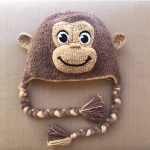 Może przedstawiać: Brązowy, dziergany na drutach kapelusz w kształcie małpy z uśmiechającą się twarzą i długimi, warkoczowymi sznurkami. Kapelusz jest wykonany z miękkiej przędzy i ma puszystą fakturę.