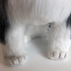 May include: Close-up of a white and black plush dog toy with black fur around the face and ears. The dog is sitting with its paws visible.