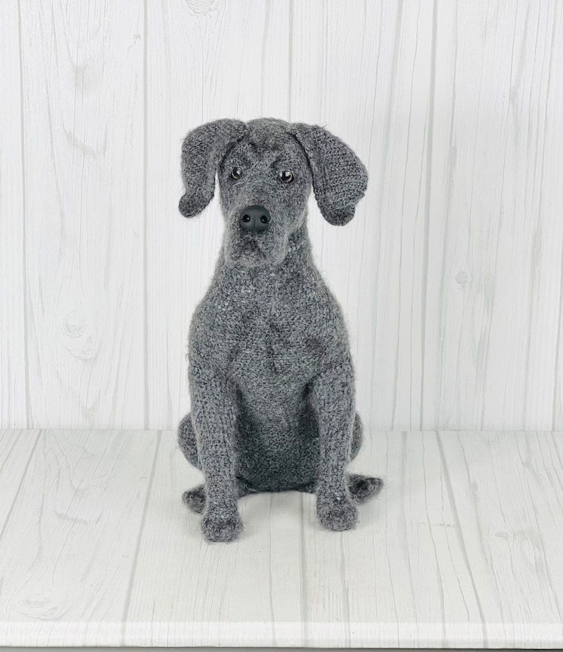 Puede incluir: Una figurita de perro de punto gris sentada sobre un fondo de madera blanca. El perro tiene un hocico largo y orejas ca&iacute;das.