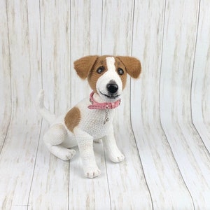 Op de afbeelding: Een gehaakt Jack Russell terrier speelgoed met bruin en wit bont, met een roze halsband met een zilveren hanger. De hond zit op een witte houten achtergrond.