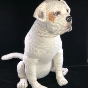 May include: White crocheted stuffed dog sitting on a black surface. The dog has brown and gray markings on its face and paws.