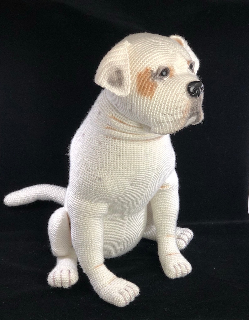 May include: White crocheted stuffed dog sitting on a black surface. The dog has brown and gray markings on its face and paws.