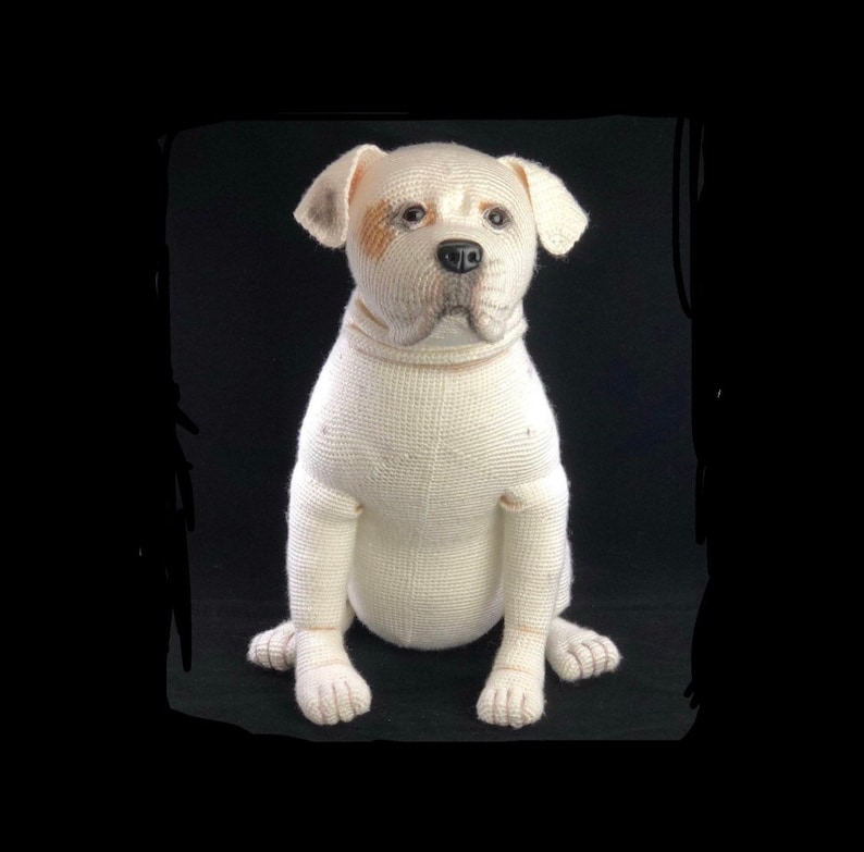 May include: A white crocheted dog with brown markings around its eyes. The dog is sitting with its paws crossed and looking directly at the camera.