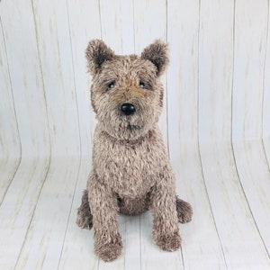 May include: A plush toy dog with brown and gray fur. The dog is sitting with its legs crossed and has a black nose and eyes.