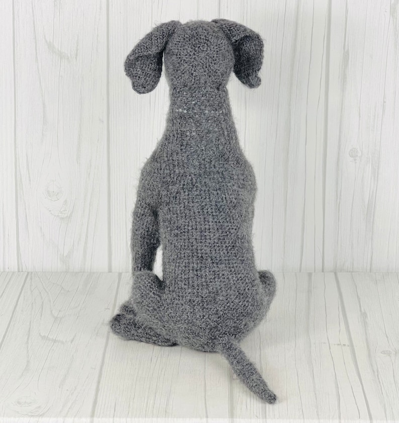 Puede incluir: Una figurita de perro de punto gris sentada sobre una superficie de madera blanca. El perro est&aacute; de espaldas a la c&aacute;mara con la cola enroscada detr&aacute;s.