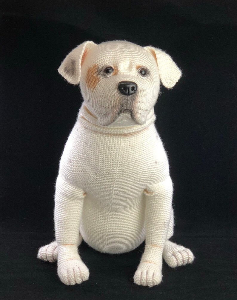 May include: White crocheted stuffed dog sitting with its paws crossed. The dog has brown and black markings around its eyes and nose.