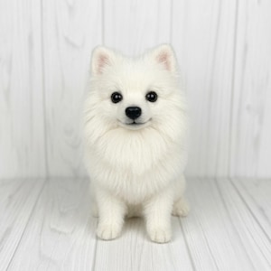 May include: A needle felted sculpture of a white Pomeranian dog with fluffy fur, upright ears, and dark, expressive eyes. The dog is in a seated position, facing forward with a black nose and a detailed, realistic facial expression.