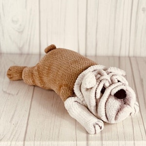 May include: A crocheted stuffed bulldog toy with brown and white fur, lying on a light wood surface.