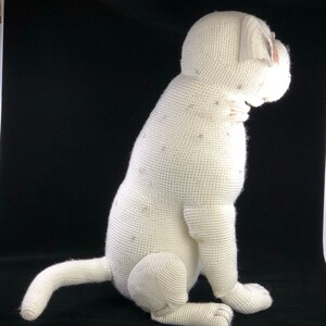 May include: White crocheted stuffed dog sitting on a black surface. The dog is facing to the right and has a white crocheted body with a brown crocheted nose.