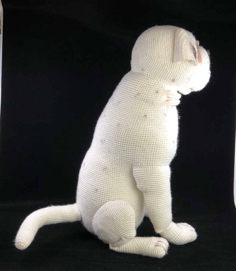 May include: White crocheted stuffed dog sitting on a black surface. The dog is facing to the right and has a white crocheted body with a brown crocheted nose.