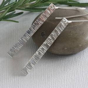 May include: A pair of silver earrings with a textured bar design. The earrings are hanging from a silver hook.