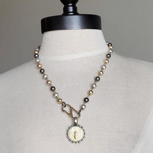 May include: A necklace featuring a strand of alternating metallic gold, silver, and dark gray spherical beads. The necklace includes a large silver-toned lobster clasp connector attached to a circular pendant with a gold-toned bee emblem.