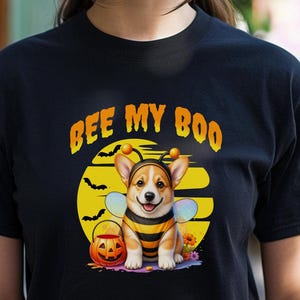 May include: A black t-shirt featuring a graphic of a Corgi dog dressed in a bee costume with wings and antennae. The design includes a yellow moon, black bats, a jack-o'-lantern bucket, and the orange text BEE MY BOO centered above the dog.