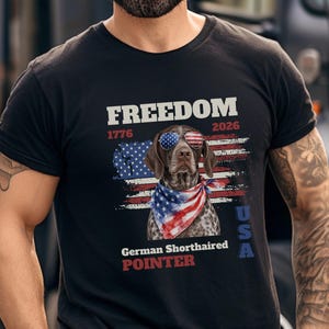 Puede incluir: Una camiseta negra con un gráfico de un braco alemán de pelo corto con gafas de sol y un pañuelo con la bandera estadounidense. El diseño incluye el texto FREEDOM, 1776, 2026, USA y German Shorthaired Pointer sobre un fondo de bandera.