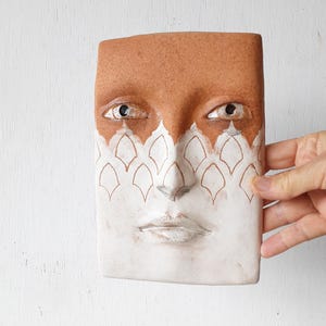 Può includere: Un'applique da parete rettangolare in ceramica raffigurante un volto stilizzato. La parte superiore è in terracotta, che sfuma nel bianco con un motivo decorativo attorno agli occhi e al naso. Occhi e labbra sono dettagliati.