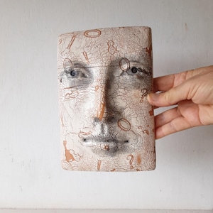 May include: A rectangular ceramic sculpture depicting a stylized face with eyes, nose, and mouth. The sculpture has a cracked, aged appearance with brown and beige tones. The surface is decorated with small, repeating symbols.
