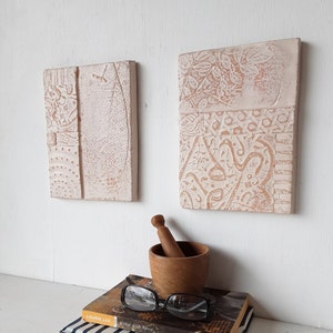 May include: Two white ceramic wall art pieces with embossed designs. The piece on the left has a geometric pattern and the piece on the right has a floral pattern. The pieces are hanging on a white wall.
