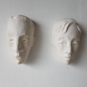 Op de afbeelding: Twee witte keramische gezichtsbeelden aan een witte muur. De gezichten hebben gesloten ogen en zijn gedetailleerd met gelaatstrekken. De sculpturen zijn waarschijnlijk bedoeld voor woondecoratie of artistieke weergave.