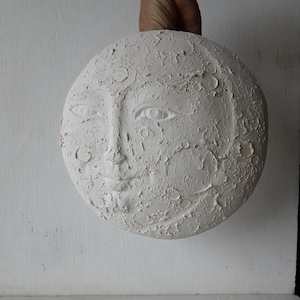 May include: A white, textured, round sculpture of a moon with a face carved into it. The moon has craters and a smiling expression. The sculpture is held by a hand.