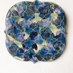 May include: A decorative ceramic tile with a quatrefoil shape featuring a repeating geometric star and cross pattern in shades of deep blue, navy, and light blue with white accents. The surface has a glossy finish with thin white grout lines.