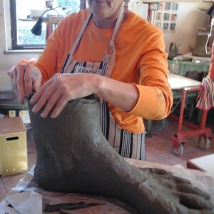 May include: A person is working on a large, gray clay foot sculpture. The foot is resting on a piece of newspaper. The person is wearing a striped apron and an orange shirt.