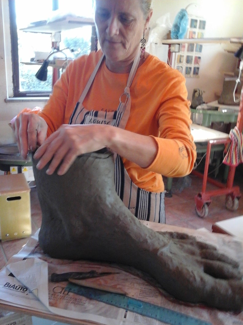 May include: A person is working on a large, gray clay foot sculpture. The foot is resting on a piece of newspaper. The person is wearing a striped apron and an orange shirt.