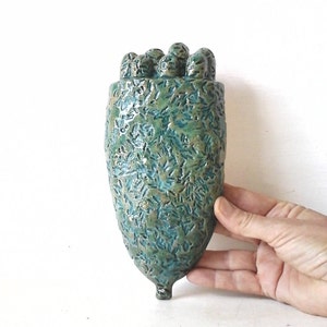 May include: A teal green ceramic wall vase with a textured surface and a unique, organic shape. The vase has a rounded top and a pointed bottom.
