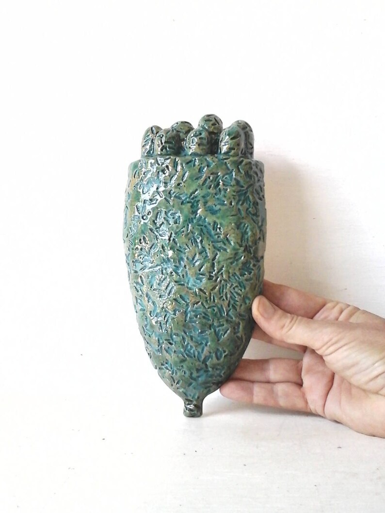 May include: A teal green ceramic wall vase with a textured surface and a unique, organic shape. The vase has a rounded top and a pointed bottom.