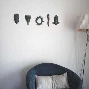 May include: A blue armchair with two white pillows sits in front of a white wall with five decorative wall hangings. The wall hangings are in a row and are all a dark green color. The wall hangings are shaped like a hand, a funnel, a sun, a long thin shape, and a fish.