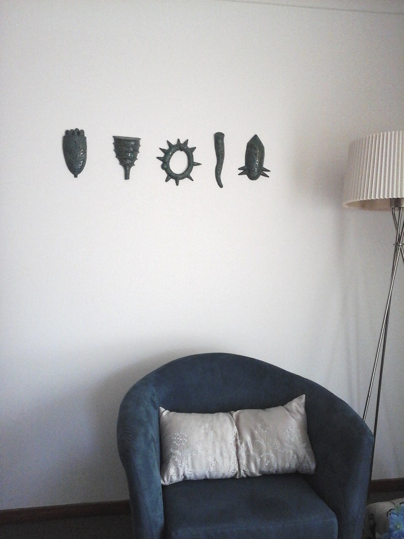 May include: A blue armchair with two white pillows sits in front of a white wall with five decorative wall hangings. The wall hangings are in a row and are all a dark green color. The wall hangings are shaped like a hand, a funnel, a sun, a long thin shape, and a fish.