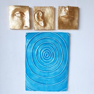 以下が含まれることがあります： 人間の目、耳、口を描いた金箔のセラミック製の壁彫刻3つが、同心円のデザインが施された青いセラミック製の壁彫刻の上に水平に配置されています。