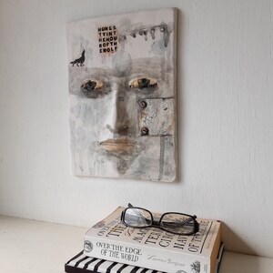 May include: A white ceramic tile with a painted face and the words "HONES TYINT HEHOU ROFTH EWOLF" on the top. The face has a neutral expression with closed lips and a small black silhouette of a wolf on the left side of the tile.