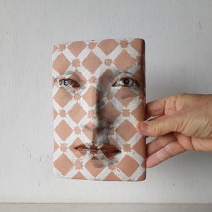 May include: A rectangular terracotta face sculpture with a diamond pattern in white and peach. The sculpture features eyes, a nose, and lips. The sculpture is held by a hand.
