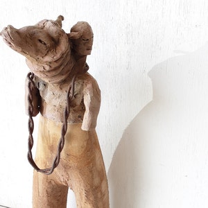 May include: A ceramic sculpture of a person with a head that resembles an animal, carrying another smaller figure on their back. The sculpture is made of brown clay and has a chain around the neck.