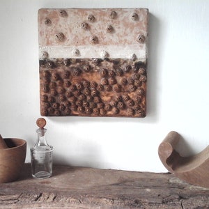May include: A square, textured artwork with a layered design in shades of brown and beige hangs on a white wall. Below, a wooden mortar and pestle, a clear glass bottle, and a carved wooden sculpture sit on a wooden surface.