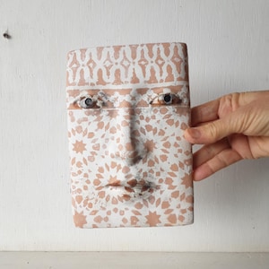 May include: A rectangular, terracotta-colored face sculpture with a white patterned design. The sculpture features a nose, lips, and eyes with metal accents. The face is held by a hand against a white background.