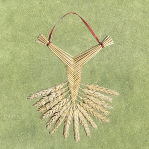 May include: A handcrafted wheat weavings ornament with a red ribbon for hanging. The ornament is made of wheat stalks and has a decorative design. The wheat is a light tan color, and the background is a muted green.