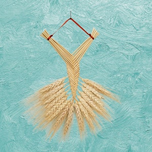 May include: A straw wheat braid woven into a dress shape, hanging from a red ribbon against a light blue textured background.