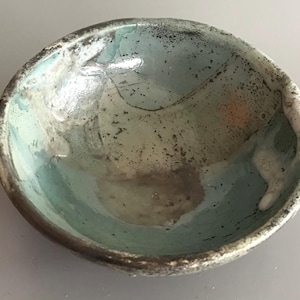 Op de afbeelding: Een handgemaakte keramische kom met een unieke, organische vorm. De binnenkant heeft een mix van blauwe, grijze en witte glazuren, wat een marmerachtig effect creëert. De rand is donkerbruin, wat contrast toevoegt. Ideaal om te serveren of als decoratief stuk.