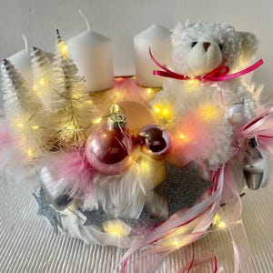 Könnte beinhalten: Ein festlicher Weihnachts-Mittelpunkt mit einem weißen Teddybär, rosa Schleife, weißen Kerzen, rosa Federn und Kugeln. Das Arrangement umfasst Miniatur-Weihnachtsbäume, Lichterketten und silberne Sternakzente, die eine warme, einladende Weihnachtsdekoration schaffen.