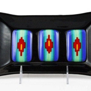 May include: A black rectangular glass dish with three colorful rectangular inserts. Each insert has a red, yellow, blue, and green striped pattern with a red cross-like design in the center. The dish is supported by clear stands.