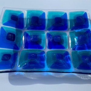 May include: A rectangular glass dish with a grid pattern of square tiles in shades of blue and teal. The tiles have a three-dimensional effect, with darker blue centers. The dish is transparent.