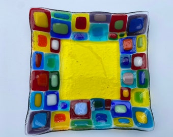 May include: A square glass dish with a yellow center and a border of colorful squares in red, blue, green, and yellow. The dish has a slightly irregular shape and is likely handmade.