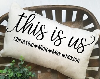 May include: A white pillow with black text that reads "This is us" with the names Christine, Nick, Max, and Mason underneath. The pillow is in a brown wicker basket.