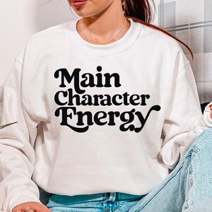 May include: White sweatshirt with the text "Main Character Energy" in a black, retro font. The person is wearing light blue jeans and sitting on a couch with patterned pillows. A book and a black and white patterned mug are also visible.
