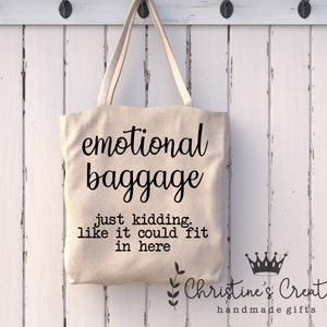 May include: A white canvas tote bag with black text that reads "emotional baggage just kidding, like it could fit in here". The bag is hanging on a white wooden hook rack. The text "Christine's Creations handmade gifts" is also visible on the bag.