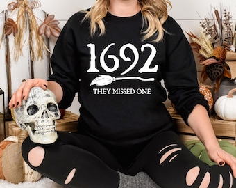 May include: Black sweatshirt with the text "1692 THEY MISSED ONE" in white. The image features a decorative skull and pumpkins, suggesting a Halloween theme. The sweatshirt is paired with black ripped leggings.